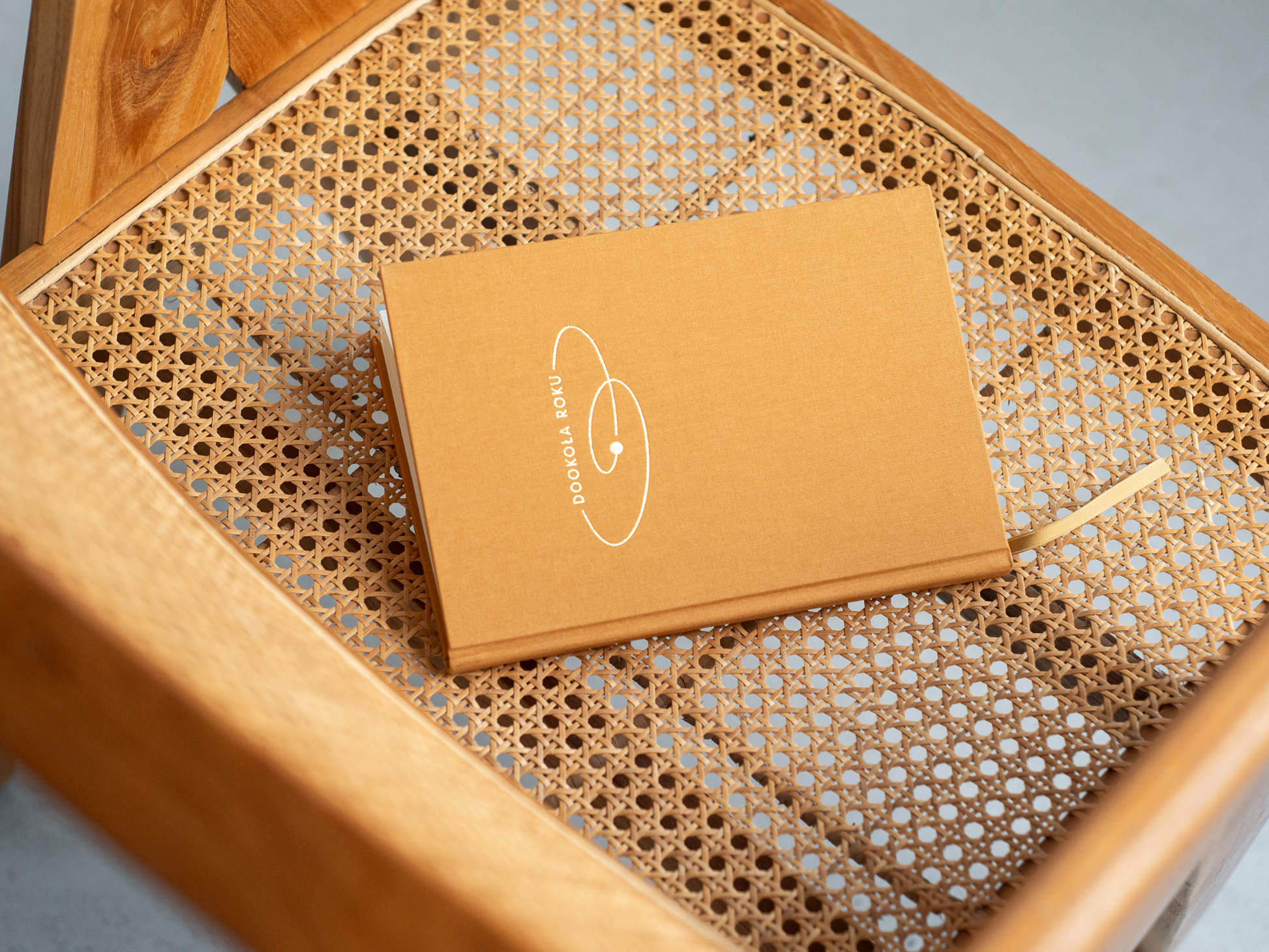 Na  zdjęciu widoczny jest zamknięty notes/planer „Dookoła Roku” w musztardowej okładce z białym minimalistycznym nadrukiem. Notes leży na siedzisku drewnianego krzesła z plecionką wiedeńską, a z jego wnętrza wystaje dopasowana tasiemka–zakładka w kolorze okładki.