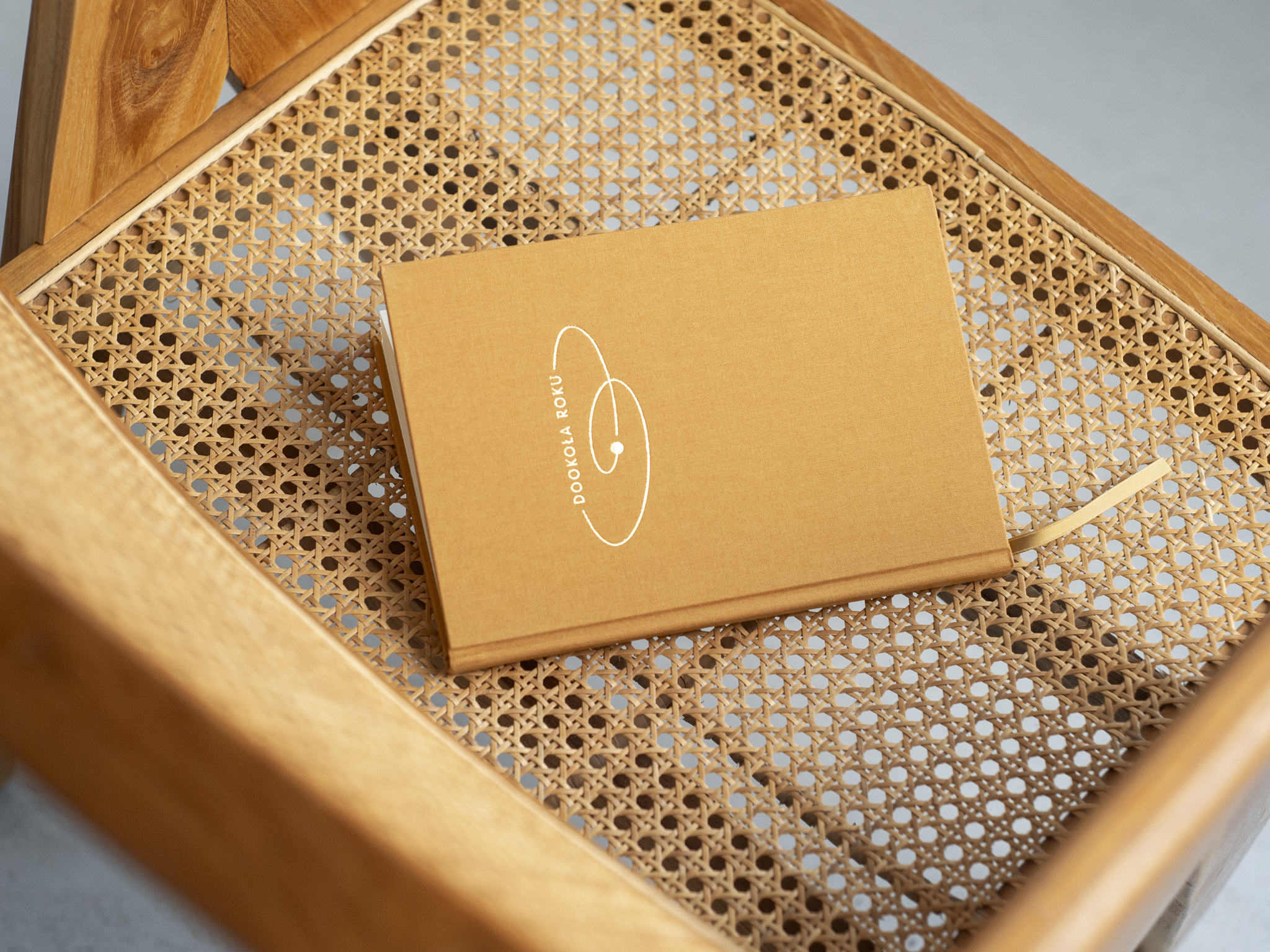 Na  zdjęciu widoczny jest zamknięty notes/planer „Dookoła Roku” w musztardowej okładce z białym minimalistycznym nadrukiem. Notes leży na siedzisku drewnianego krzesła z plecionką wiedeńską, a z jego wnętrza wystaje dopasowana tasiemka–zakładka w kolorze okładki.