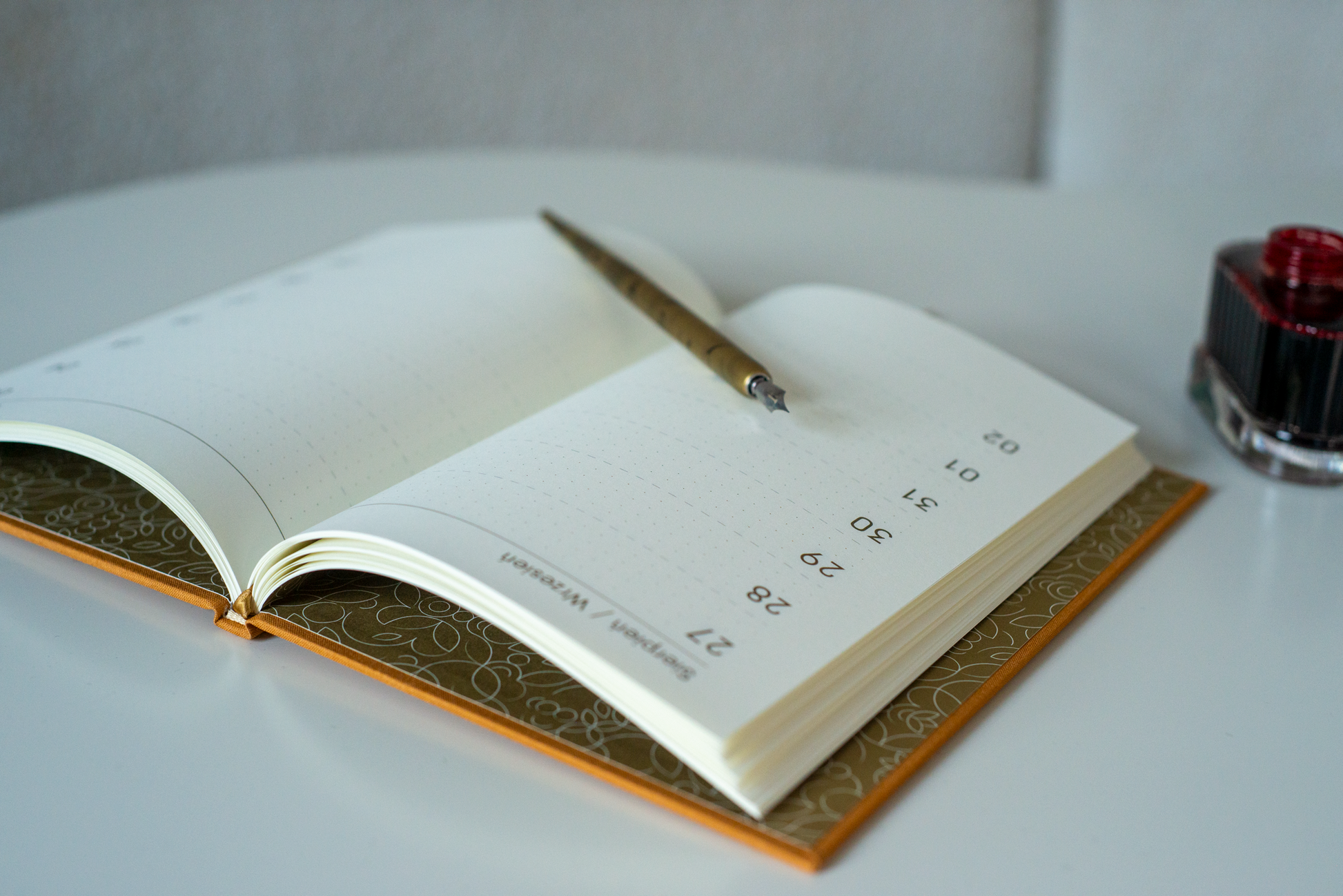 Na zdjęciu widoczny jest otwarty planer „Dookoła Roku” z rozkładem dni od 27 sierpnia do 2 września. Notes ma jasne strony w kropki i ozdobną wyklejkę w roślinne wzory. Na jego środku leży złote pióro, a obok, na blacie stołu, stoi kałamarz z czerwonym atramentem.