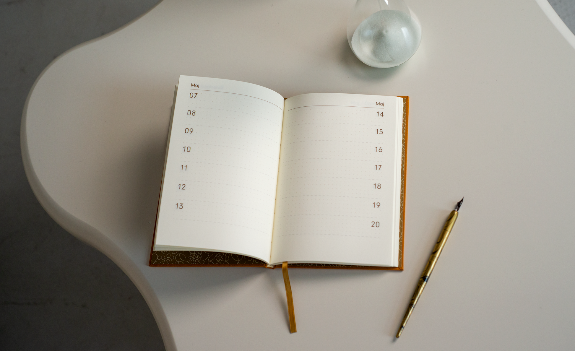 Na zdjęciu widoczny jest otwarty notes/planer „Dookoła Roku” z widocznymi datami od 7 do 20 maja. Notes leży na jasnym, falistym blacie stołu, obok znajduje się klepsydra z jasnym piaskiem oraz złote pióro.