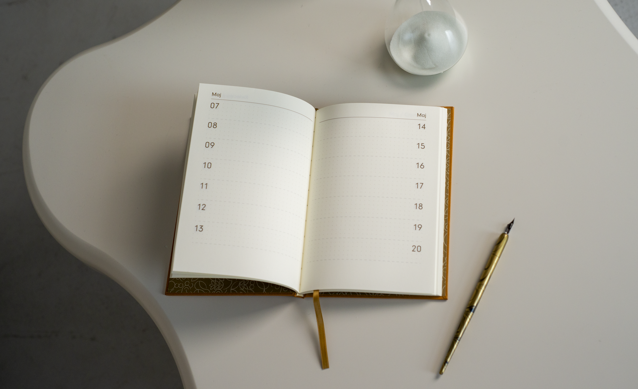 Na zdjęciu widoczny jest otwarty notes/planer „Dookoła Roku” z widocznymi datami od 7 do 20 maja. Notes leży na jasnym, falistym blacie stołu, obok znajduje się klepsydra z jasnym piaskiem oraz złote pióro.