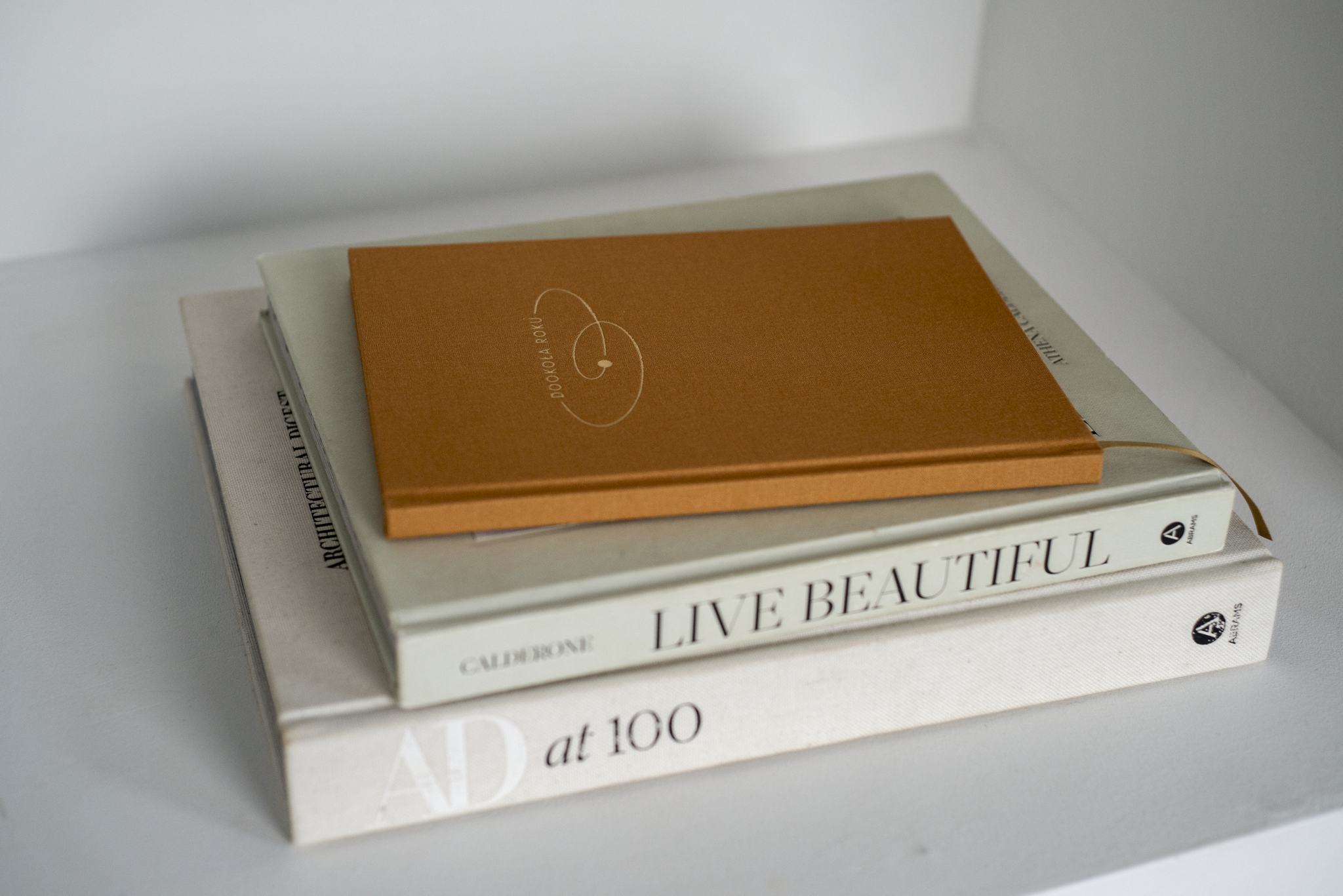 Na zdjęciu widoczny jest zamknięty notes „Dookoła Roku” w musztardowej okładce z białym minimalistycznym nadrukiem. Notes leży na stosie eleganckich albumów i książek o designie i architekturze, m.in. „Live Beautiful” oraz „AD at 100”, co tworzy estetyczną, wnętrzarską aranżację.