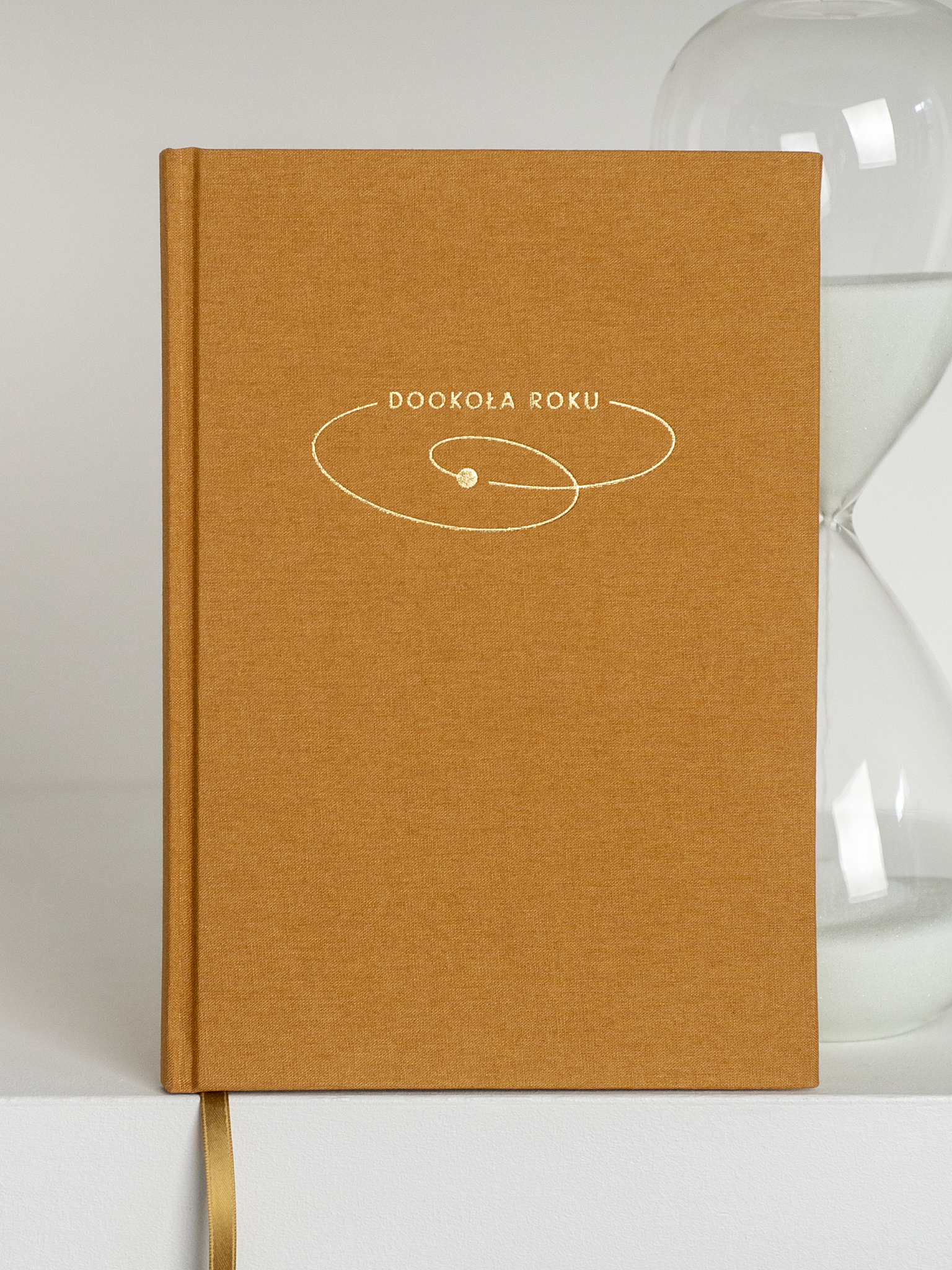 Na zdjęciu widoczny jest zamknięty notes „Dookoła Roku” w musztardowej okładce z białym minimalistycznym nadrukiem w formie linii i kropki. Notes stoi pionowo na jasnej powierzchni, a obok niego znajduje się szklana klepsydra z jasnym piaskiem. Z dolnej części wystaje dopasowana tasiemka–zakładka.